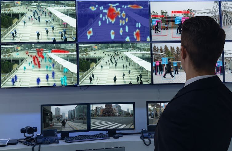 Man in suit watches screens showing surveillance camera footage