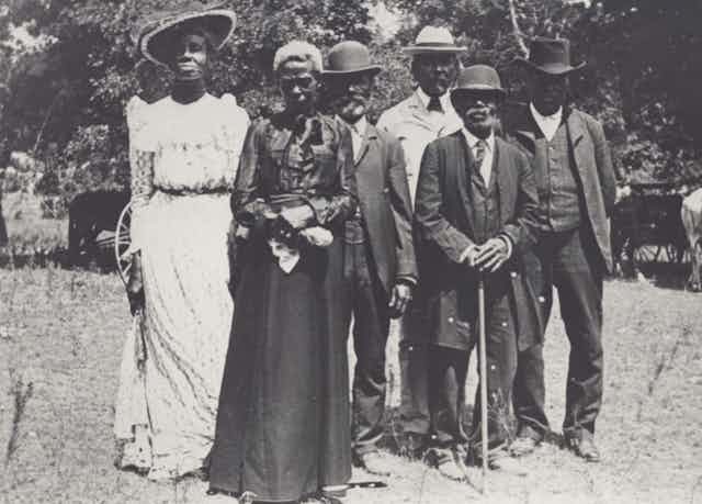 In this black-and-white image taken in the early 1900s, a group of Black men and women are wearing business suits and fancy dresses to celebrate Emancipation Day.dressed up in suits 