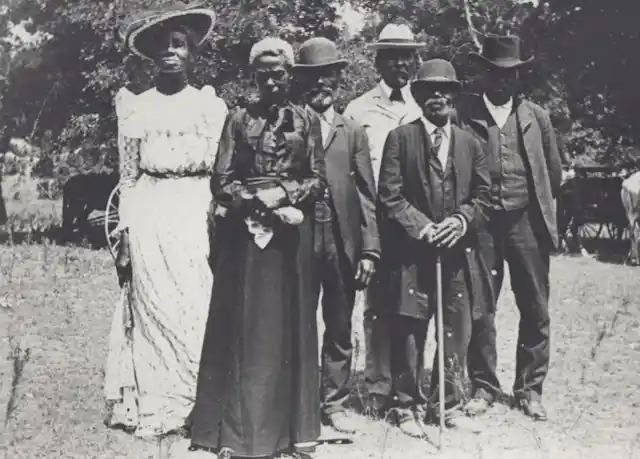 In this black-and-white image taken in the early 1900s, a group of Black men and women are wearing business suits and fancy dresses to celebrate Emancipation Day.dressed up in suits