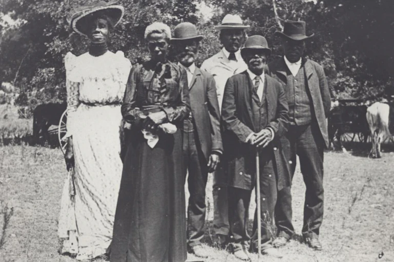 In this black-and-white image taken in the early 1900s, a group of Black men and women are wearing business suits and fancy dresses to celebrate Emancipation Day.dressed up in suits
