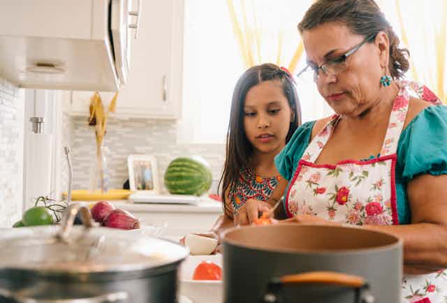 A Latina grandmother and granddaughter stand side by side in the kitchen, peering at notes, with pots of food, fruits and vegetables on the counter in front of them.