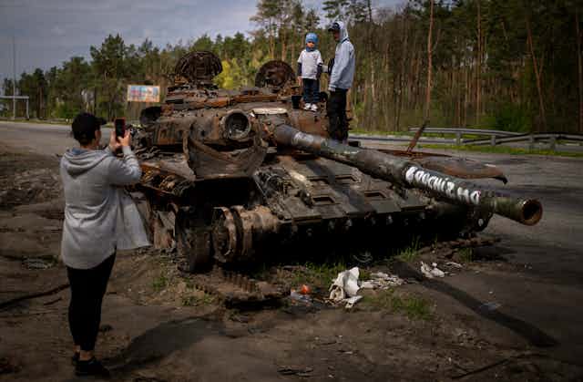 Two young boys stand atop a destroyed Russian tank as a woman takes their picture