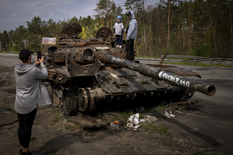 Two young boys stand atop a destroyed Russian tank as a woman takes their picture