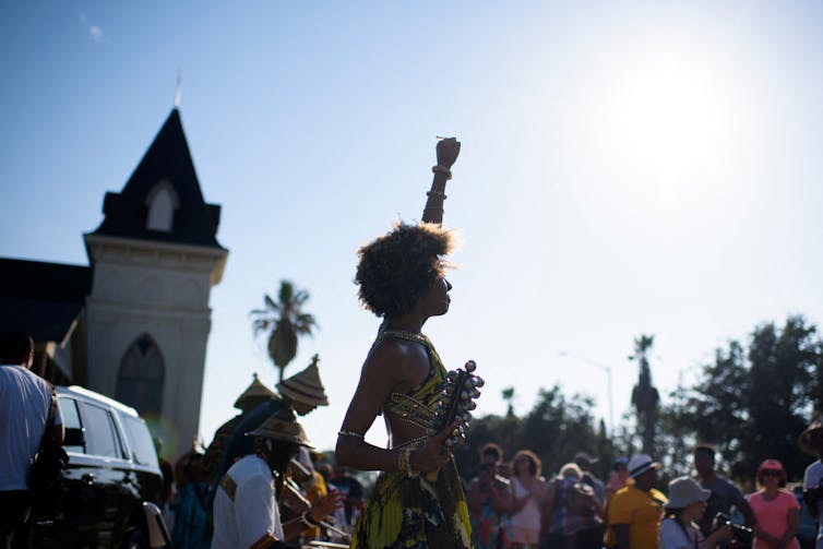 With a blue sky in the background, a Black woman stands over a crowd of people, raising her fist in the air.