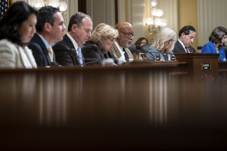 A group of men and women, looking down at notes as they sit at a high table, all in a row.