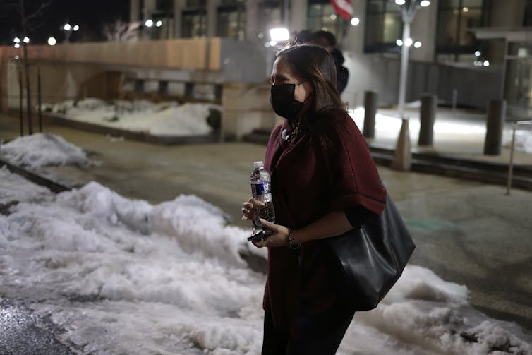 A dark-haired woman carrying a water bottle and with a bag slung over her should, leaves a building and walks on snow-covered pavement..