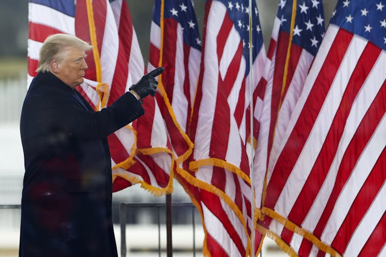 Former President Donald Trump in a black coat, standing in front of many American flags, pointing his gloved finger at something.
