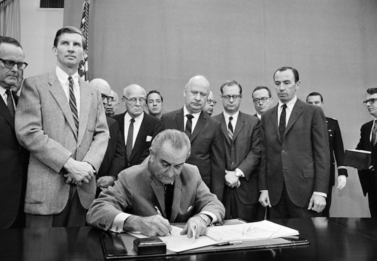 In the foreground, a balding white man signs a document. A group of middle-aged men in suits stand in the background, looking on.