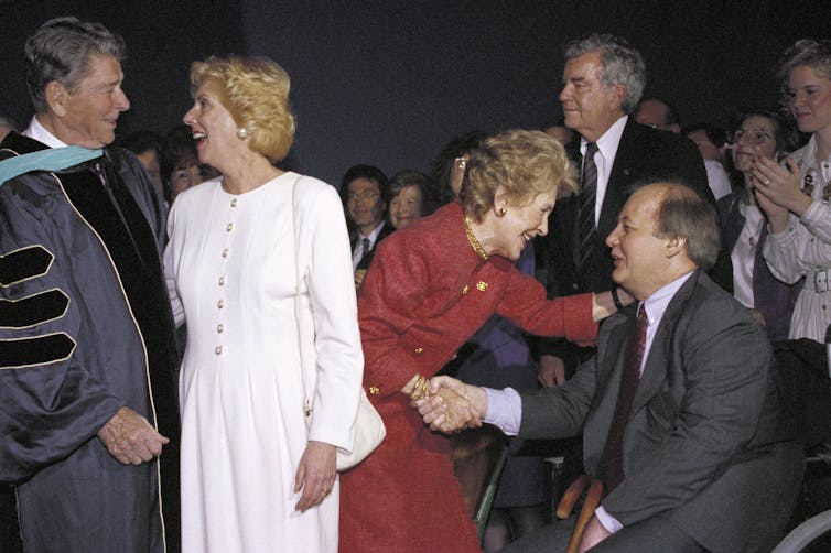 An older couple shakes hands with a man in a wheelchair and a woman in a white dress.
