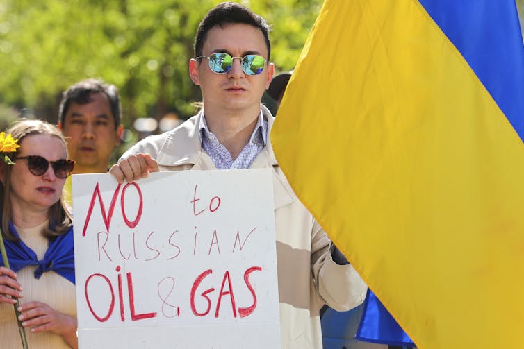 A man in sunglasses carries a Ukrainian flag and a sign that reads No to Russian Oil and Gas.