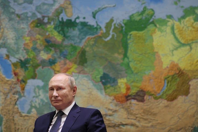 A balding man is seen with a giant map of the world behind him.