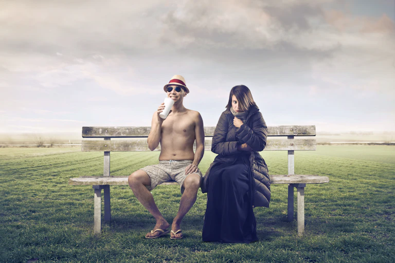 Man sitting on bench in shorts and no shirt, woman rugged up in big coat