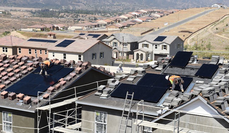 Workers prepare solar-paneled roofs on two new homes in a neighborhood with other homes with solar roofs in behind them.