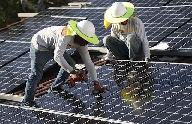Two workers install solar panels on a tile roof. One holds a drill. Both  wear sun hats with wide brims.