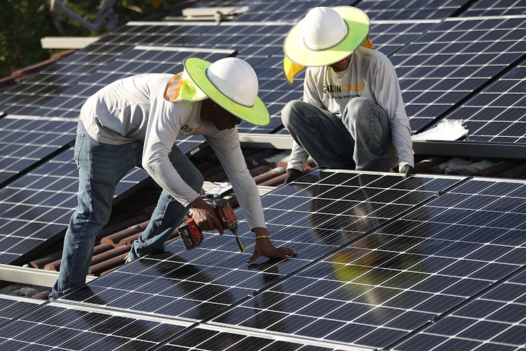 Two workers install solar panels on a tile roof. One holds a drill. Both wear sun hats with wide brims.