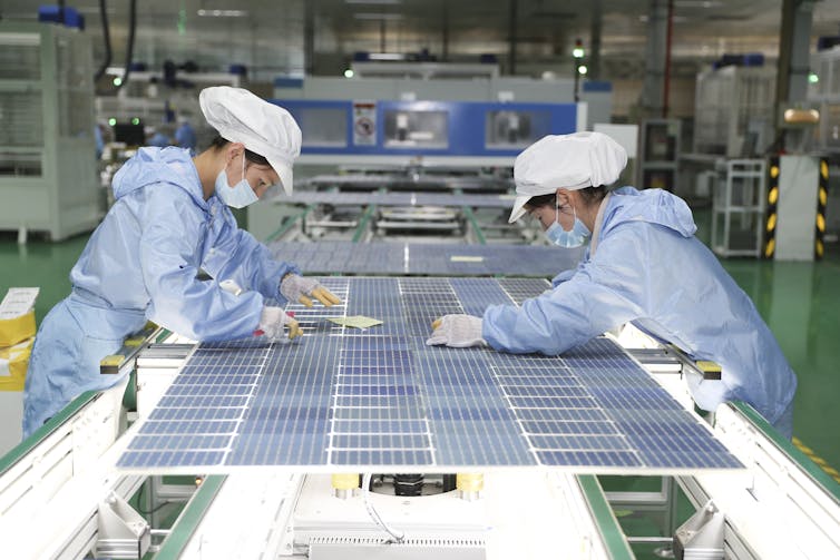 Two women in clean-suits, caps and gloves bend over a table with solar modules.