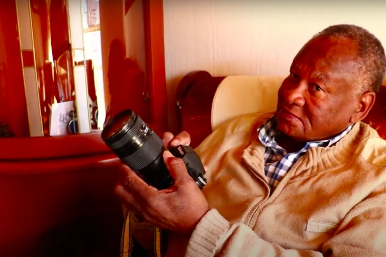 An older man sits in a chair holding a camera in his hands and looking up.