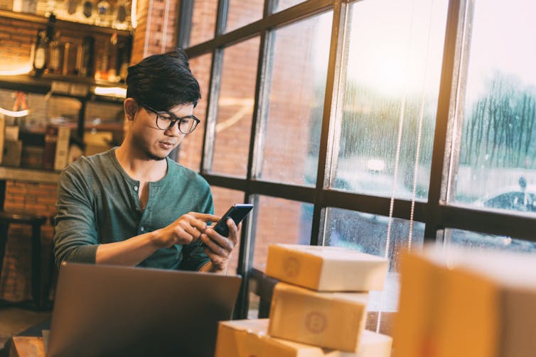 Young man in a store peruses his phone, with a laptop open on a table in front of him