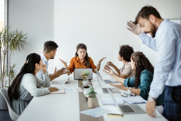A businesswoman in an orange shirt sitting at the head of the table during a meeting, with four co-workers seated around her and one standing.