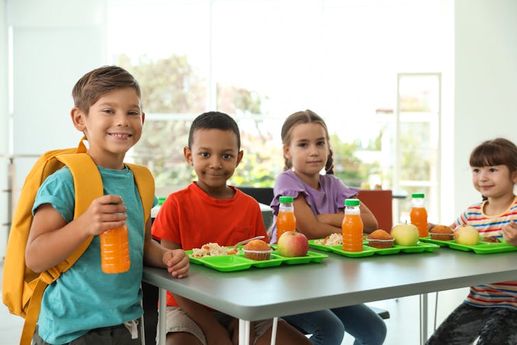 Four children, three of them sitting at a table with green lunch trays.