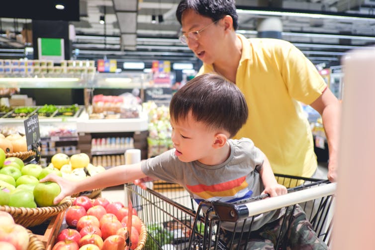 A child sitting in a grocery cart reaching for produce, with a man in a yellow shirt behind him