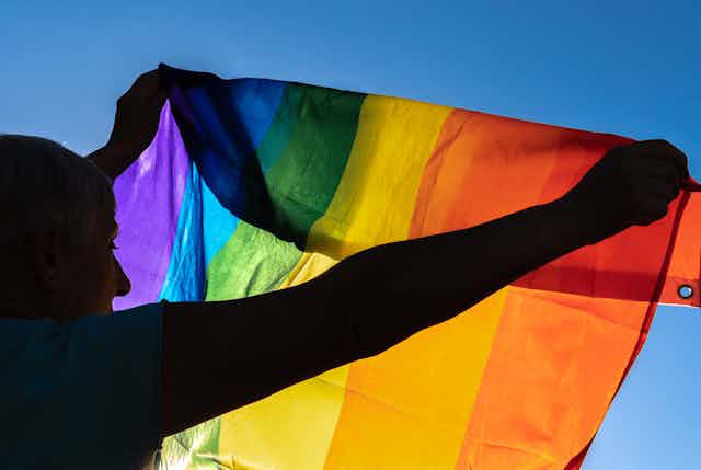 A backlit photo of an older woman holds the pride flag to the sky