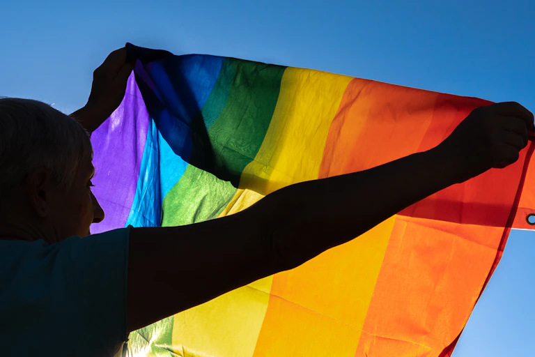 A backlit photo of an older woman holds the pride flag to the sky