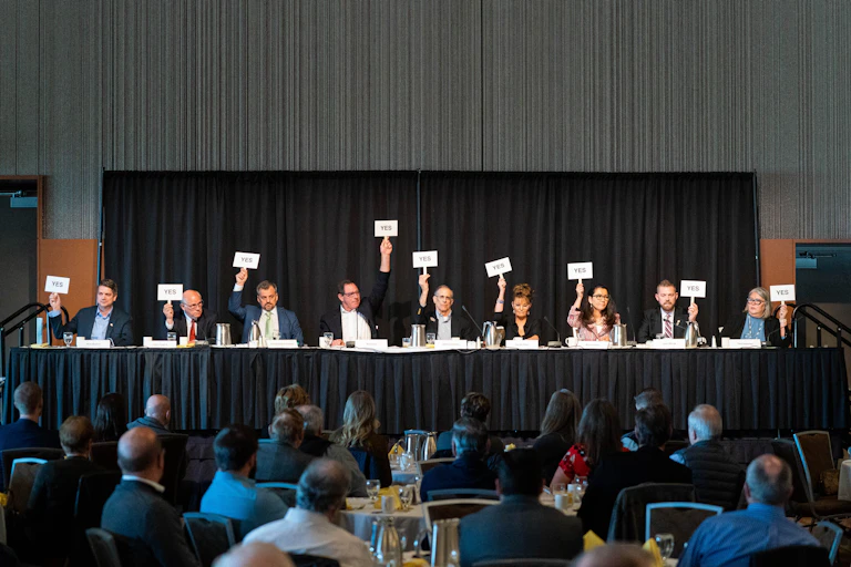 Nine people at a long table on a stage hold up white cards.