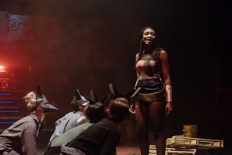 Actors in character seen on a stage: In the right of the frame, standing, appears to be a cis-gendered Black woman wearing a bronze structured tube-top-type bustier and long metal-style wristbands, surrounded by crouching figures who appear to be actors wearing animal masks.