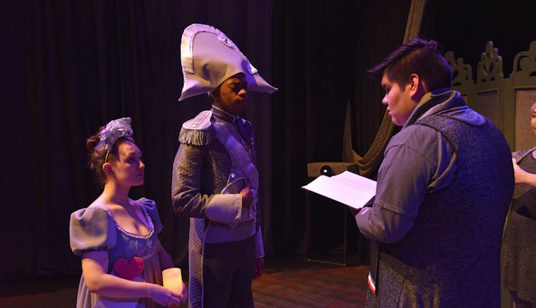 Three actors in character seen on a stage: one appears to be a cis-gendered white woman in a fancy dress, the second appears to be Black man in dignitary-soldier-type 19th century uniform with shoulder epaulettes and the third appears to be an Indigenous man in a dark coat reading from a piece of paper.