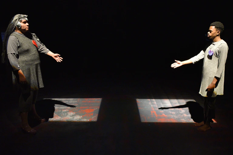 Two actors in character seen on a stage: They wear tunic-style draped clothing, with hearts pinned to their chests, and stand to the far right and far left of the frame, and are illuminated in light squares on a dark stage; the figure on the right of the frame is Indigenous and the figure on the right is Black, and they extend their hands towards one another.