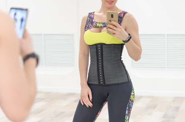 Woman in athletic clothes wearing a waist trainer takes a selfie in the mirror with her phone.
