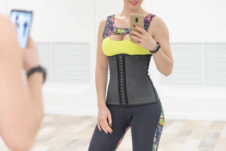Woman in athletic clothes wearing a waist trainer takes a selfie in the mirror with her phone.