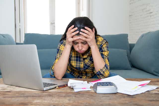 A woman in a plaid shirt sits in front of a desk with papers, a laptop, a calculator and holds her head in her hands.