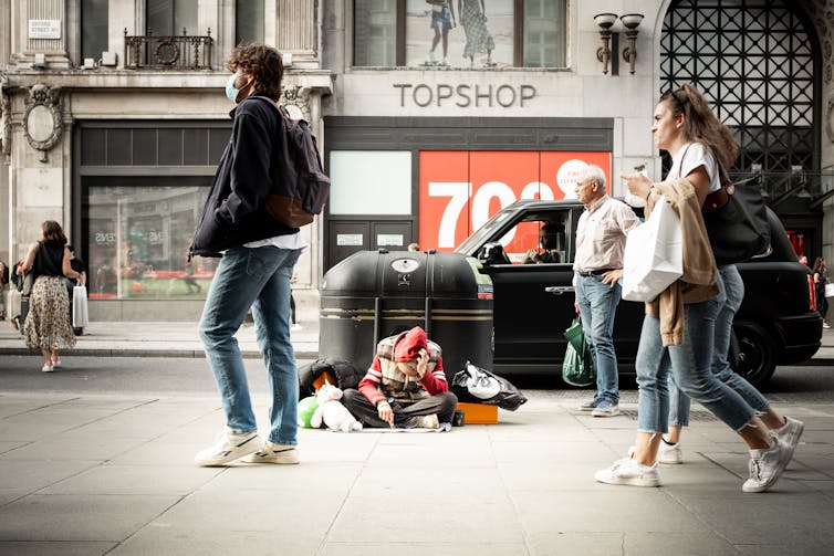 A homeless person sits on the pavement of a shopping street.