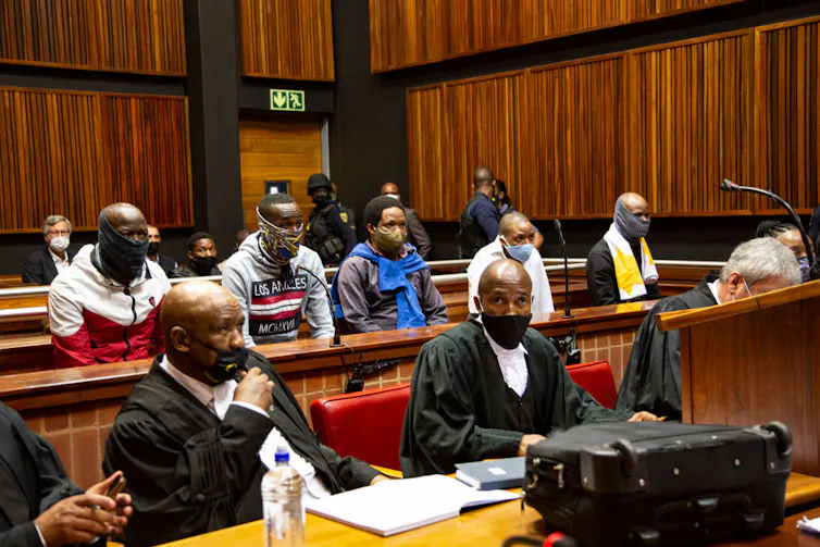 A wood-panelled room with court officials sitting in front of a row of men and people watching further back, everyone wearing face masks.