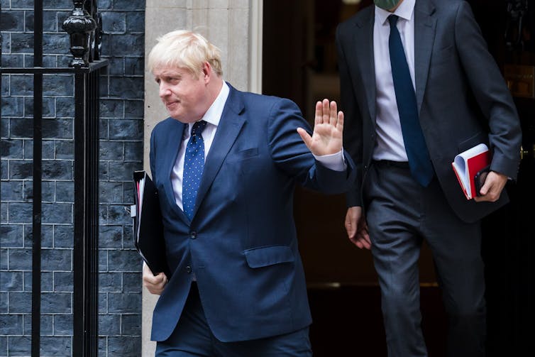 Boris Johnson walks out of Downing Street with one hand raised in greeting, looking away from the camera