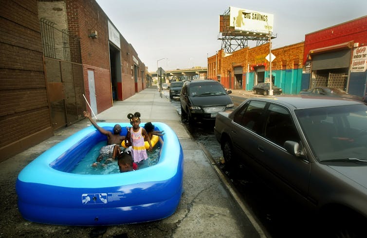 Four children in an inflatable pool on a sidewalk in an area of ​​brick buildings, cement, sidewalk and no trees.