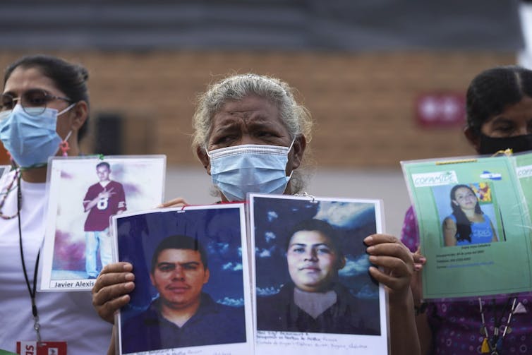 Three women, all in masks, hold up photos of their loved ones.