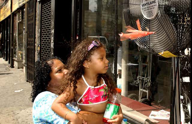 A woman holds her young daughter up to the fan on a street with storefronts and lots of pavement.