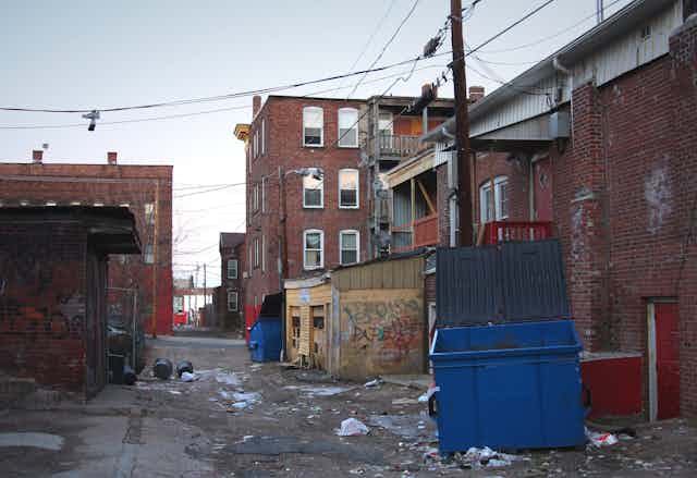 A trash-filled urban alleyway in an inner city urban neighborhood, with graffiti, trash and poorly maintained, aging buildings in the background.