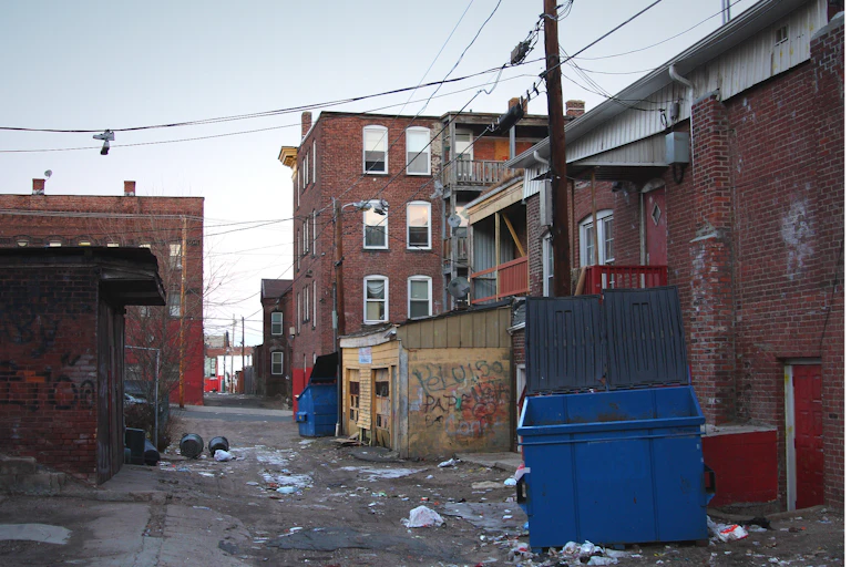 A trash-filled urban alleyway in an inner city urban neighborhood, with graffiti, trash and poorly maintained, aging buildings in the background.