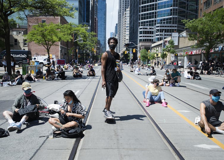 A Black man stands on a skateboard among people sitting on a city street during a protest