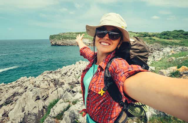 A smiling woman backpacker takes a selfie in front of a rocky coastline