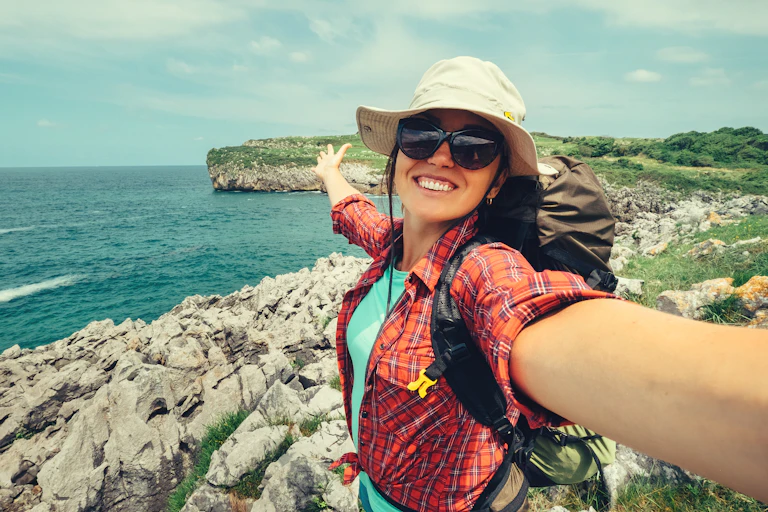 A smiling woman backpacker takes a selfie in front of a rocky coastline