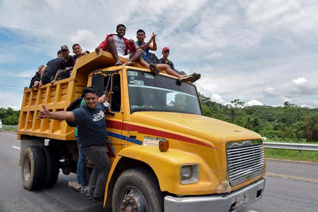 Men sit on top of a yellow truck and hang off its sides, with one giving a thumbs up