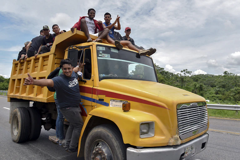 Men sit on top of a yellow truck and hang off its sides, with one giving a thumbs up