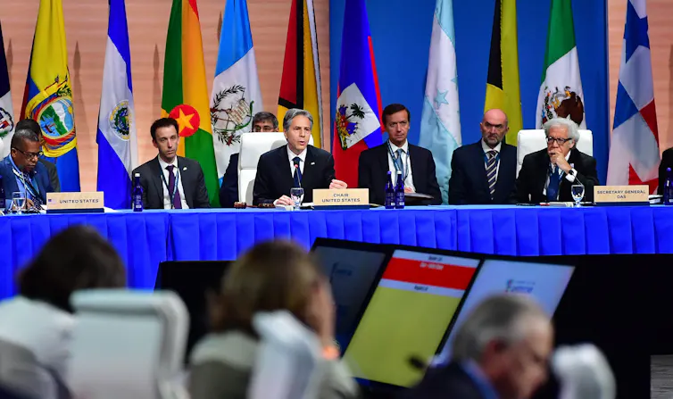 Antony Blinken is pictured at a long table with other men in suits, sitting in front of flags from Latin America.