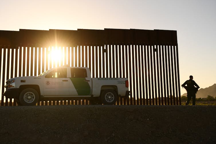 A Customs and Border Protection pick up truck is shown in front of the US-Mexico border wall. A man stands next to the wall.