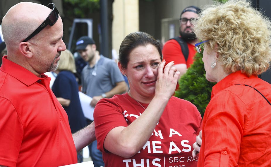 A woman dressed in a red T-shirt wipes her tears as her two friends standing next to her comfort her.